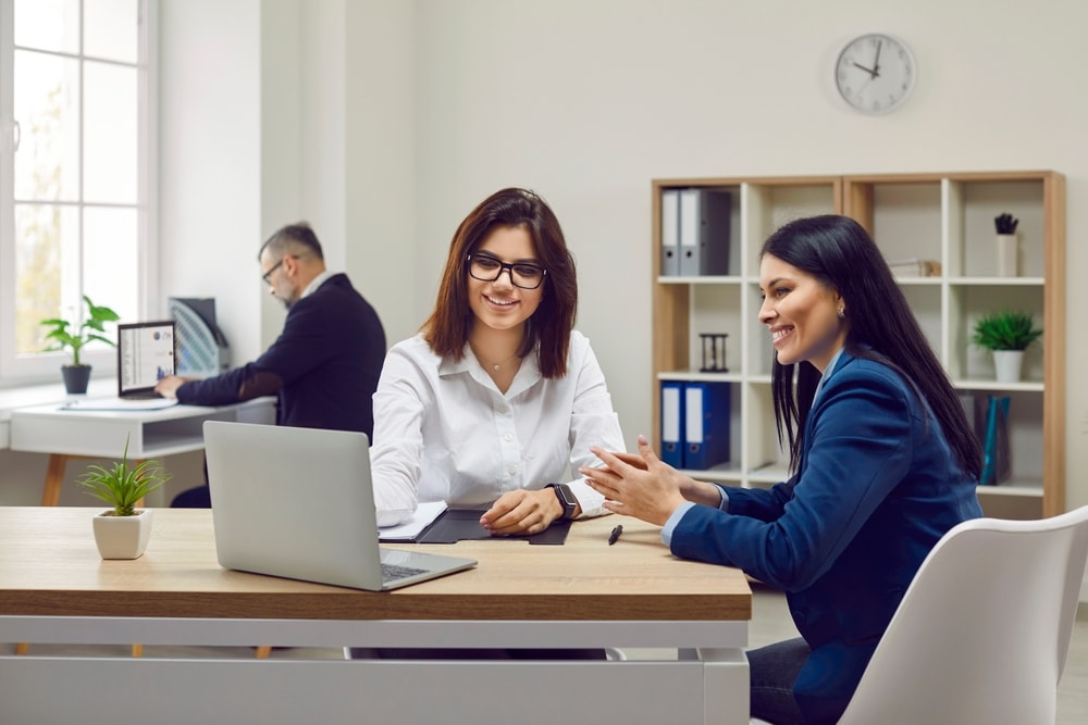 Two young women smiling discussing policies with laptop on table in front of them